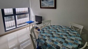 Una habitación con una mesa redonda cubierta con un mantel a cuadros, dos sillas blancas, un televisor sobre una mesita y un cuadro enmarcado en la pared.