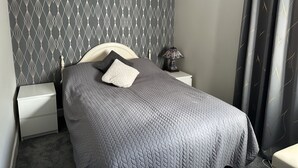 A neatly made bed with a grey quilt, a bedside lamp, and a patterned wallpaper.