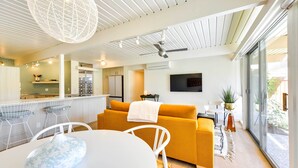 A modern kitchen with white cabinetry, a dining area with a white table and chairs, and a living area with a yellow sofa and a TV mounted on the wall.