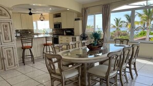 Una cocina amplia con una isla central, un comedor con mesa y sillas redondas, y ventanas grandes que ofrecen vistas a palmeras y al océano.