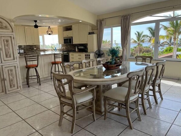 Una cocina amplia con una isla central, un comedor con mesa y sillas redondas, y ventanas grandes que ofrecen vistas a palmeras y al océano.