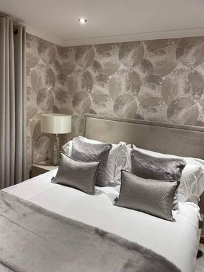 A neatly made bed with a grey blanket and pillows, a headboard, a bedside lamp, and a patterned wall. - Remarkable 2-bed Apartment in Auchterarder (Auchterarder)