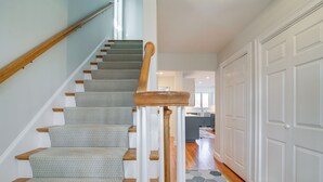 A staircase with wooden steps and carpeted landings, leading to a kitchen area with a large island and white cabinetry. - Spacious home for entertaining, centrally located to beach, ferry, and airport. (Edgartown)