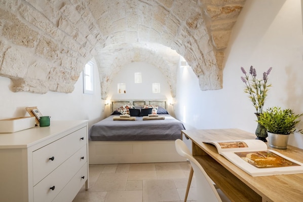 A bedroom with a stone ceiling, a bed with pillows and a blanket, a bedside table with a book, a dresser, and a potted plant.