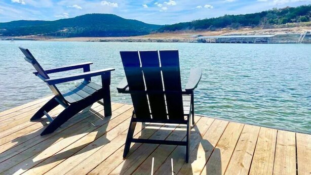 A wooden deck with two Adirondack chairs facing a calm lake, surrounded by hills and a clear sky.