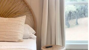 A bed with a woven headboard, a round wicker side table, and a view of trees through a window.