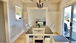 A dining area with a long table, white chairs, a chandelier, and a view of a balcony with outdoor seating. - Amazing Views and Walkable to Downtown! Historic 3/2 Home, Sleeps 8 (Tiburon)
