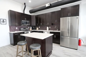 A modern kitchen with dark cabinets, a stainless steel refrigerator, a microwave, and a large island with stools.