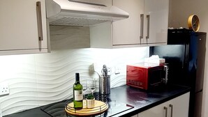 A modern kitchen with white cabinets, a black countertop, and a built-in oven. There is a wine bottle and glasses on the counter, and a red appliance in the background. - Maple House - Inviting 1-bed Apartment in London (London)