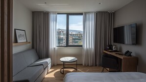Una habitación de hotel moderna con cama, sofá, una mesita, escritorio con silla, televisión y ventana con cortinas.