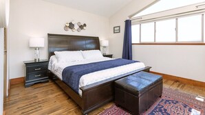 A neatly arranged bedroom with a large bed, bedside tables, a decorative wall piece, and a view of the outdoors through a window. - Two-Bed Cabin – Breathtaking Fjord View at the Summit House. (Malahat)