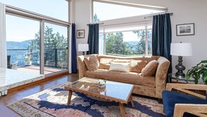 A living room with a sofa, a glass coffee table, a patterned rug, and a view of a balcony with a railing and potted plants. - Two-Bed Cabin – Breathtaking Fjord View at the Summit House. (Malahat)