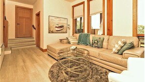 A living room with a beige sofa, a glass coffee table, and a painting of a boat on the wall. - Panoramic Suite  – Breathtaking Fjord View, Summit House. (Malahat)