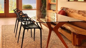 A modern interior with a glass dining table, black chairs, a wooden sofa, and large windows offering a view of mountains. - Panoramic Suite  – Breathtaking Fjord View, Summit House. (Malahat)
