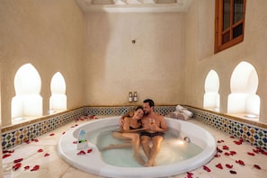 Una pareja en una bañera con pétalos de rosa en el suelo, un termómetro y una ventana con vistas.