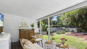 Un espacio de estar al aire libre cubierto con mobiliario de mimbre, una pequeña mesa con una bebida y flores, y vistas a un jardín exuberante.