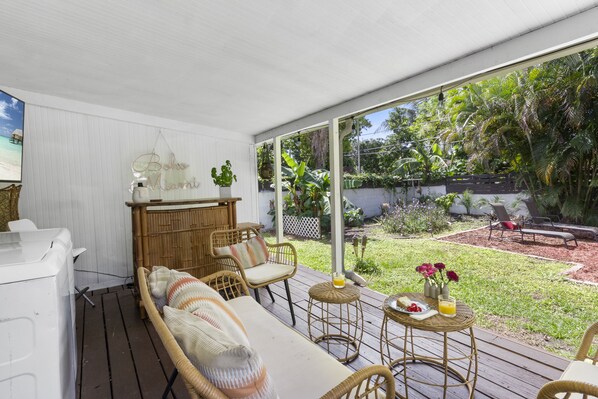 A covered outdoor seating area with wicker furniture, a small table with a drink and flowers, and a view of a lush garden.