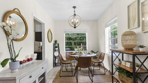 Un comedor con una mesa redonda, sillas, una araña y una mesa de buffet con una cesta y plantas.