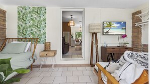 Habitación luminosa con cama, mesita de noche, espejo, lámpara, televisor y vista a una playa a través de la ventana.