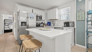 A modern kitchen with white cabinets, a stainless steel refrigerator, a microwave, and a coffee maker. There is a small island with two stools and a colorful plate. - DOG-FRIENDLY, OUTDOOR POOL, DUPLEX BEACH HOUSE!! (Gulf Shores)