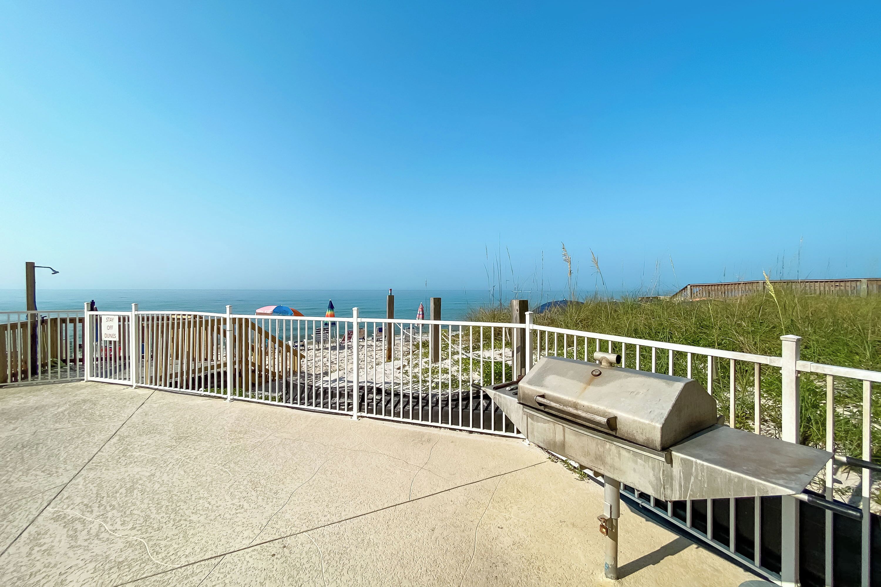 Une terrasse au bord de la plage, avec un barbecue, une clôture blanche et une vue sur l’océan.