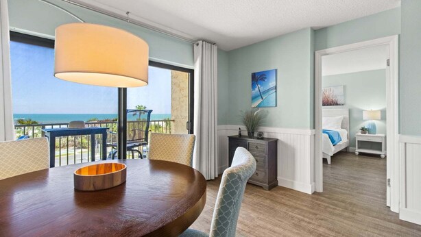 Une chambre d’hôtel équipée d’une table à manger, de chaises, offrant une vue sur l’océan, avec un lit, une table de chevet et un tableau au mur.