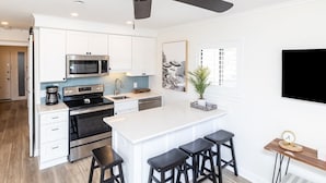 A modern kitchen with white cabinetry, a central island with bar stools, a ceiling fan, and a mounted flat-screen TV. - Guest Favorite~Steps to the beach~Pickleball and Tennis Courts~Beach Bar (Hilton Head Island)