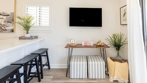 A modern living room with a flat-screen TV, a wooden table with stools, a striped ottoman, and a potted plant. - Guest Favorite~Steps to the beach~Pickleball and Tennis Courts~Beach Bar (Hilton Head Island)