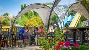An outdoor seating area with wooden tables and chairs, surrounded by a wooden deck and covered by a canopy structure. - Camping familial 4* | Mobil-home + Accès piscine (Monterblanc)