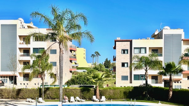 Ein Poolbereich mit Liegestühlen, Palmen und modernen Apartmentgebäuden im Hintergrund.
