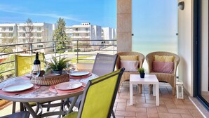 Ein Balkon mit Tisch für zwei Personen, Korbstühle und Blick auf das Meer und Gebäude im Hintergrund.