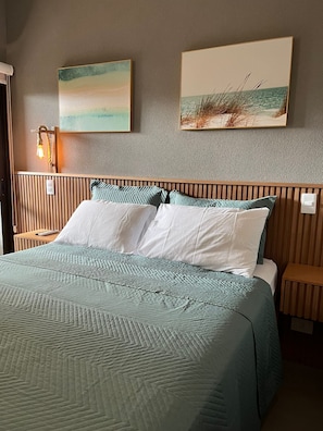 A neatly made bed with a patterned bedspread, two pillows, and a headboard with vertical slats. There are two bedside tables with lamps, and two framed pictures on the wall above the bed.