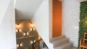 A staircase with a wooden door at the top, a wall with a geometric light fixture, and a green leafy wall to the right. - Palulu Flats (São Sebastião)
