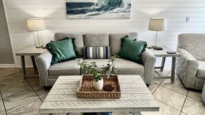 Un salon avec un canapé gris, deux tables d’appoint avec des lampes, une table basse avec une plante et un plateau, et un tableau représentant une vague, accroché au mur.