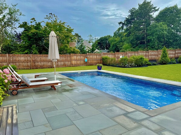 A swimming pool with a tiled deck, lounge chairs, and a wooden fence in the background. - Katama home with pool and on bike path! Walk to Edgartown and South Beach! (Edgartown)