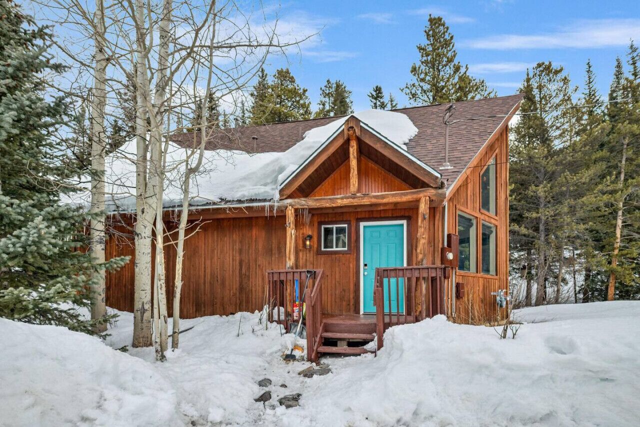 Una cabaña de madera con puerta azul, rodeada de nieve y árboles.