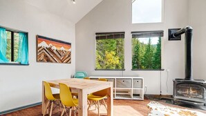 Una sala de estar moderna con una mesa de comedor de madera, sillas amarillas, una chimenea y ventanas grandes con vistas al bosque.