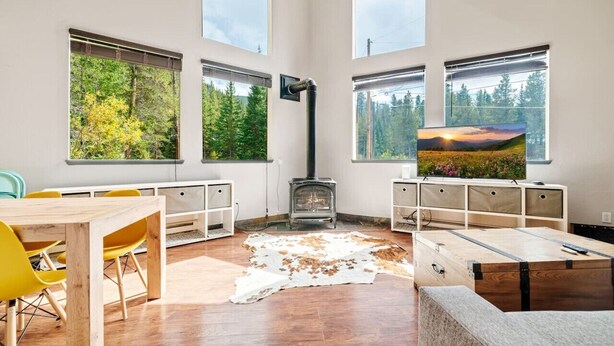 Una sala de estar moderna con una estufa de leña, ventanas amplias con vistas al bosque, un comedor con sillas amarillas y un televisor de pantalla plana grande sobre un mueble de madera.