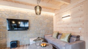 Un salon chaleureux avec un mur en pierre, un canapé gris, une table ronde sur laquelle sont disposés des fruits et un téléviseur fixé au mur.