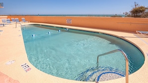 Una piscina trasparente con un cartello di bagnino, sedie a sdraio e vista sulla spiaggia.