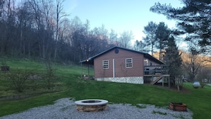 Une cabane rustique avec un foyer en pierre en premier plan, entourée d’une zone recouverte de gravier et d’une colline herbeuse.