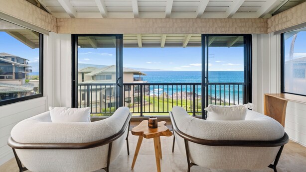 Een balkon met twee witte fauteuils, een klein houten tafeltje en uitzicht op zee en huizen.