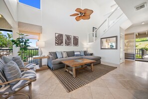 A spacious living area with a sofa, coffee table, and striped cushions. There is a large window with a view of greenery outside.