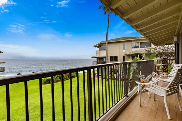 A balcony with a view of the ocean, a palm tree, and a dining area with chairs and a table. - 2bd endless luxury, and soothing sounds of waves (Lahaina)