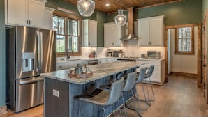 A modern kitchen with stainless steel appliances, a central island with bar stools, and a herringbone tile backsplash. - Whiskey Hollow Lodge - Mountain Views | Outdoor Living | Hot Tub (Morganton)