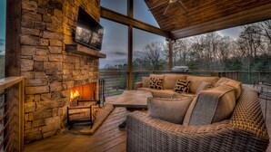 A stone fireplace with a fire, a wicker sofa with cushions, and a wooden deck with a railing. - Whiskey Hollow Lodge - Mountain Views | Outdoor Living | Hot Tub (Morganton)