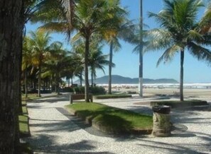 A coastal walkway lined with palm trees and a bench, leading to a beach with a view of the ocean and distant mountains. - Hotel Pousada PG One  (Praia Grande)
