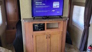 A compact RV interior with a flat-screen TV mounted on a wooden cabinet, a small seating area, and a kitchenette. - The Budget Suite (Leadwood)