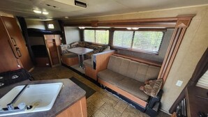 A compact RV interior with a kitchenette, a dining area, and a seating area with a window. - The Budget Suite (Leadwood)