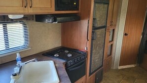 A compact RV kitchen with wooden cabinets, a stove, a microwave, and a sink. - The Budget Suite (Leadwood)
